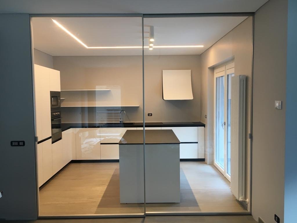 Cucina moderna con armadi bianchi e piano di lavoro nero, isola centrale, elettrodomestici integrati, grande finestra a doppia anta con tende bianche, pareti grigie, pavimento in legno chiaro, illuminazione a soffitto con faretti LED