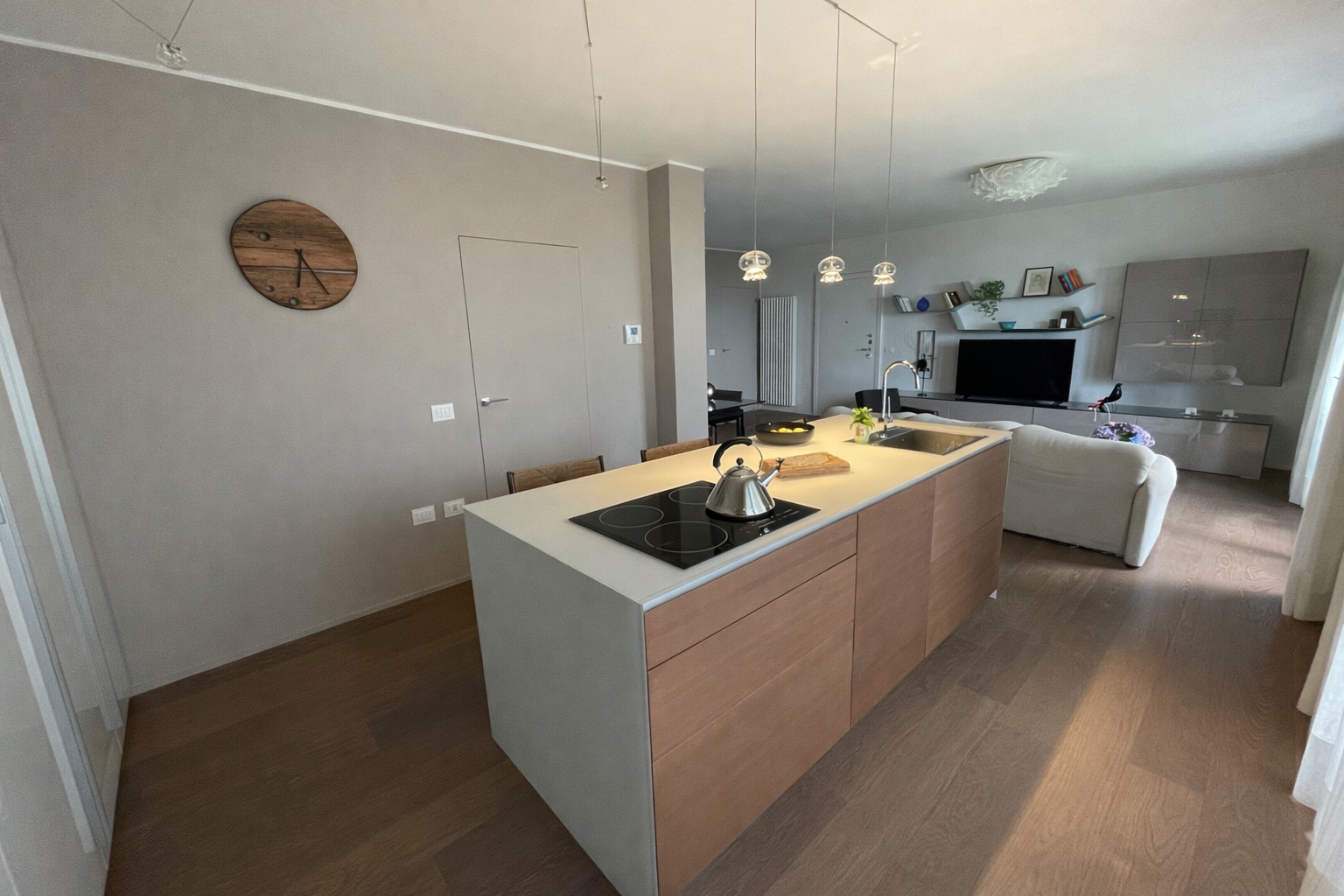 Ambiente di una cucina moderna e soggiorno con mobili in stile minimalista, pareti beige, TV e librerie sospese, divano bianco, soffitto con illuminazione sospesa e pareti decorate con quadri e piante.