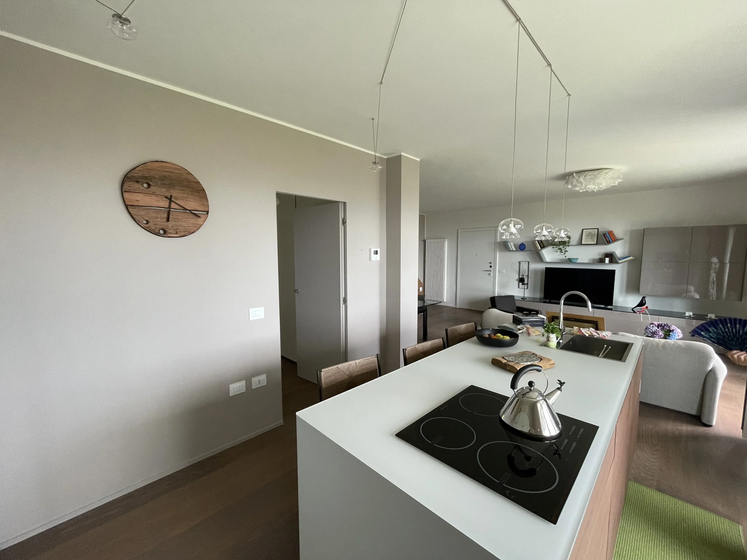 Interno di un appartamento con cucina a vista e soggiorno. Visible un tavolo da cucina con piano cottura, presenza di utensili e decorazioni, e un ambiente luminoso con divano e televisione.