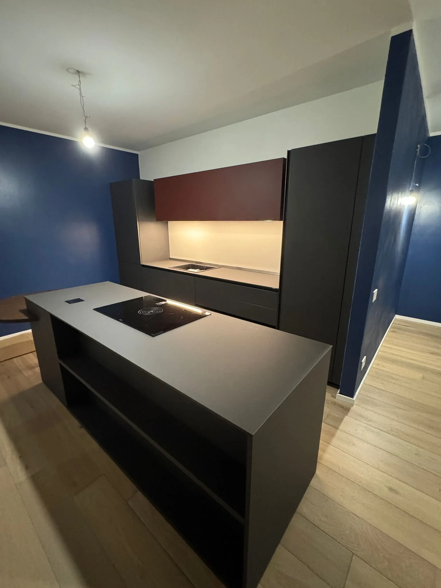 Cucina moderna con mobili neri, pensile color borgogna, pareti blu e pavimento in legno chiaro.