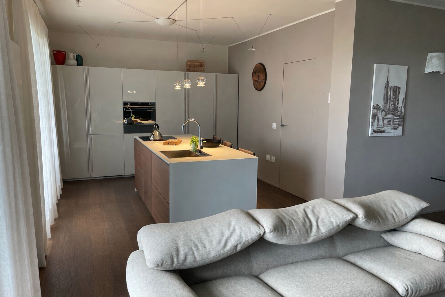 Interno di una moderna casa con area soggiorno e cucina a vista, divano beige e pareti decorate con quadri, con una cucina bianca e tavolo centrale in primo piano.