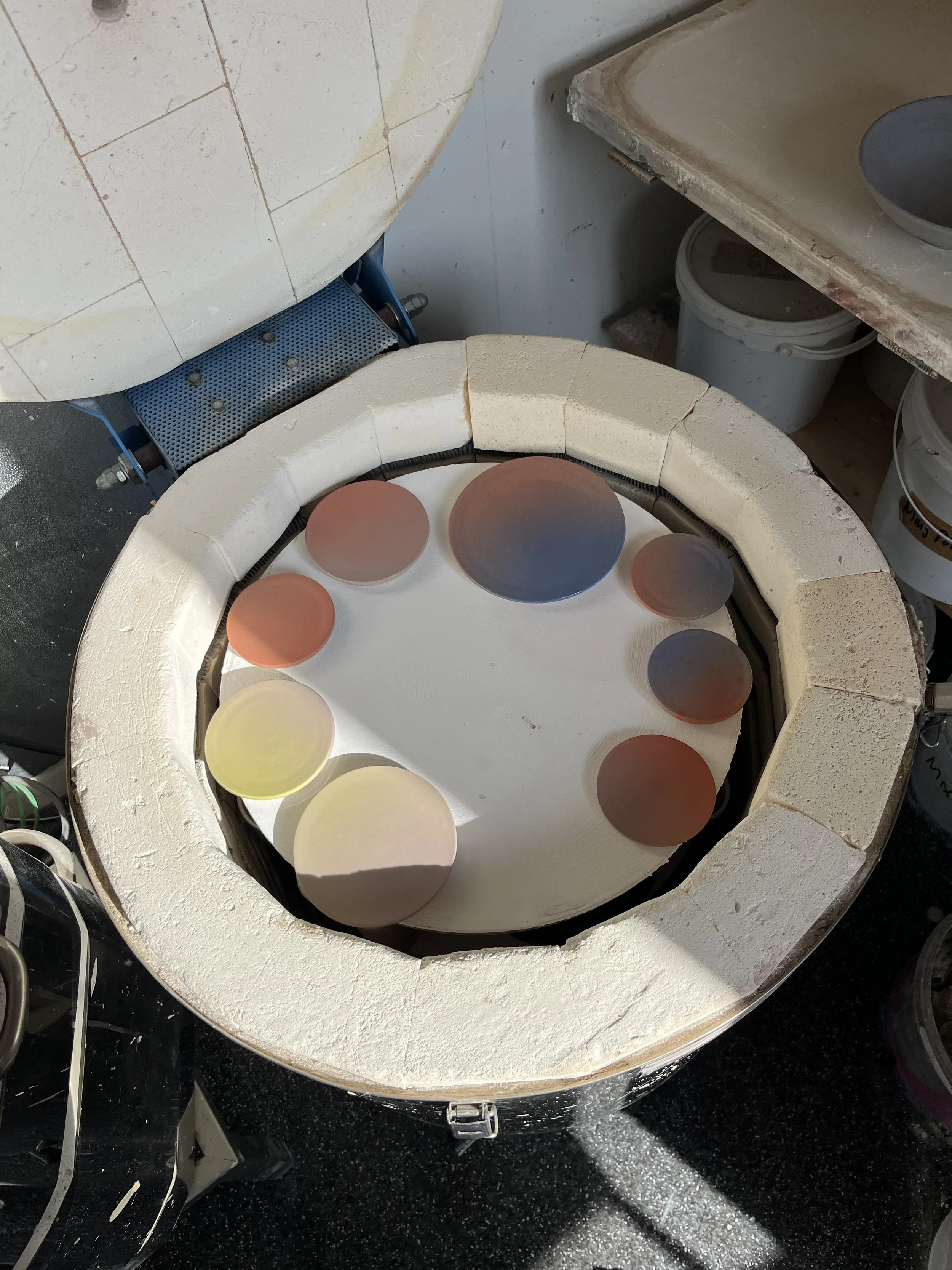 Blick in einen Keramikofen mit mehreren farbigen, glasierten Keramikstücken der verschiedenen Größen. Der Ofen ist offen und die Keramikstücke sind auf einer weißen Scheibe im Inneren des Ofens platziert.