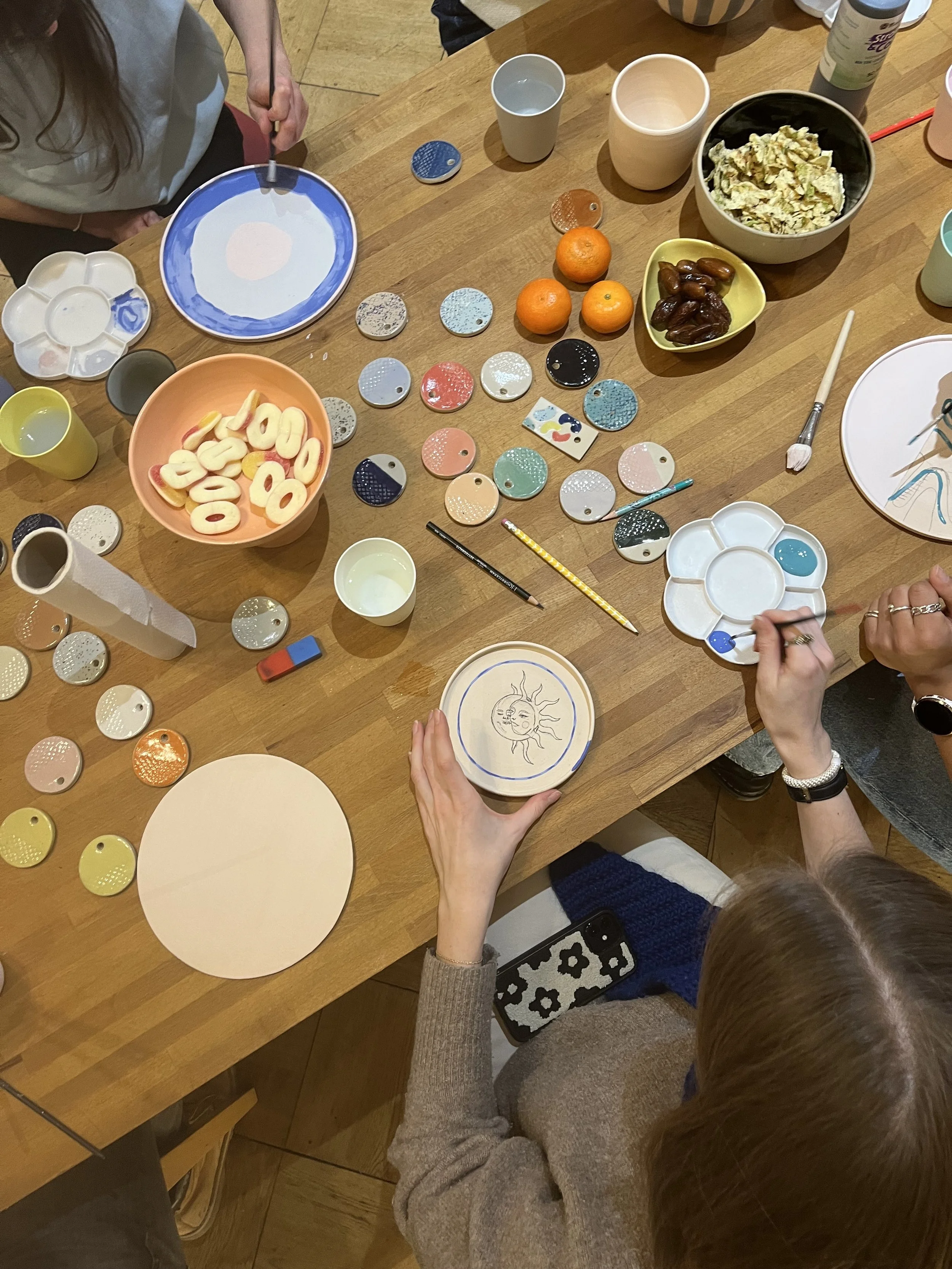 Menschen bemalen und verzieren Keramikgegenstände bei einer Bastelaktivität auf einem Holztisch, umgeben von verschiedenen bunten Farben, Pinseln, Snacks und Getränken.