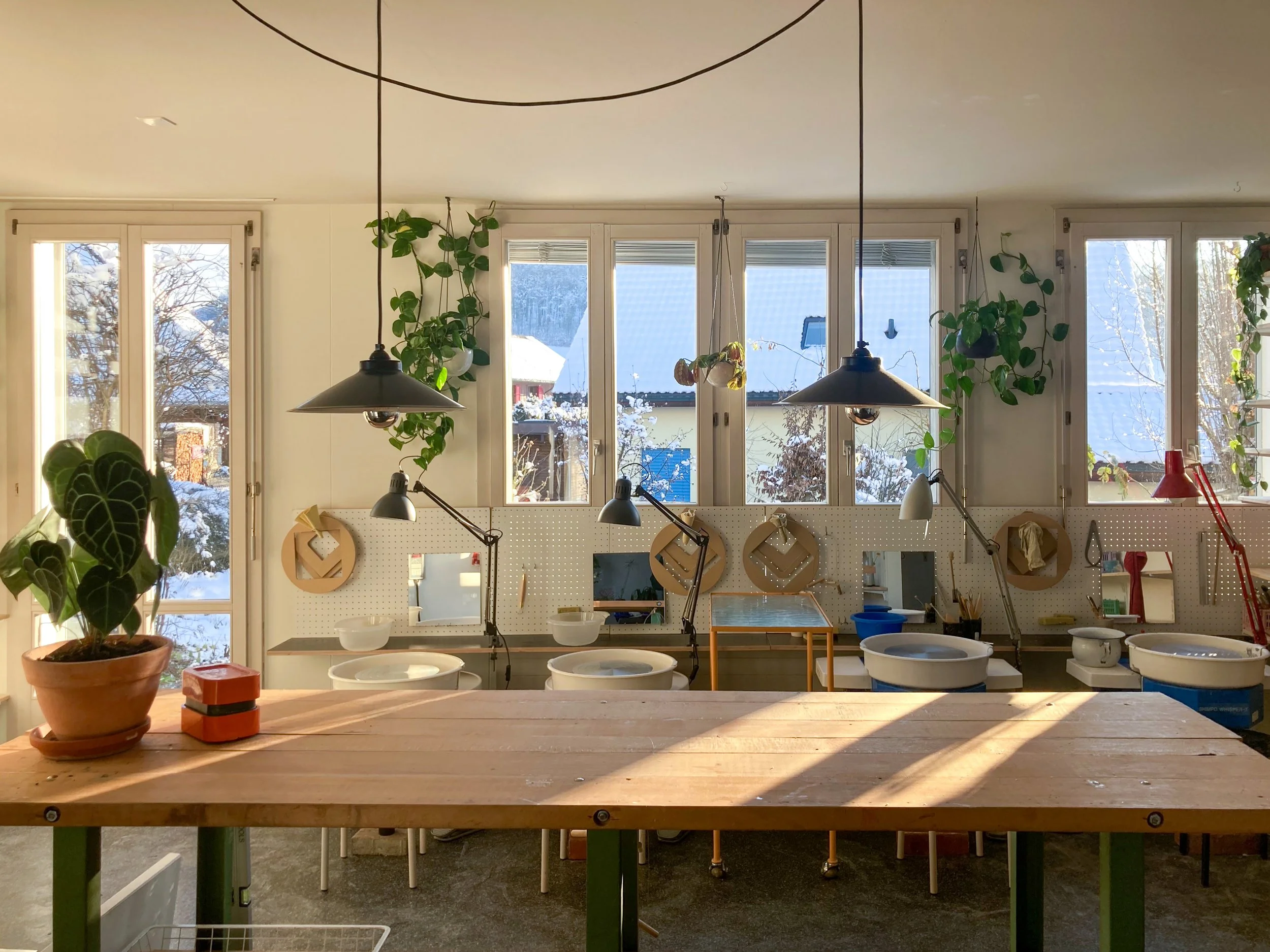 Ein hell erleuchteter Werkraum mit großen Fenstern, Pflanzen an den Wänden und Arbeitsstationen mit Tischen und Töpfen, vermutlich ein Keramik- oder Töpferstudio.