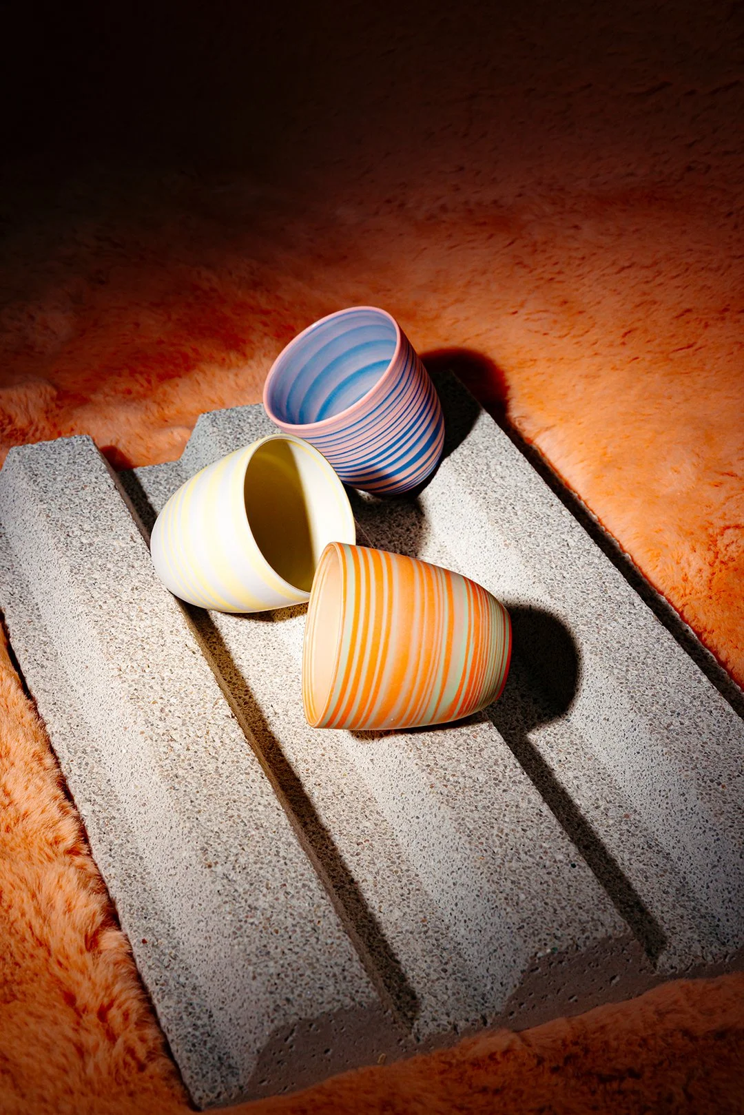 Drei bunte, gestreifte Keramikbecher auf grauem Ziegelstein, umgeben von orangefarbenem, flauschigem Stoff.