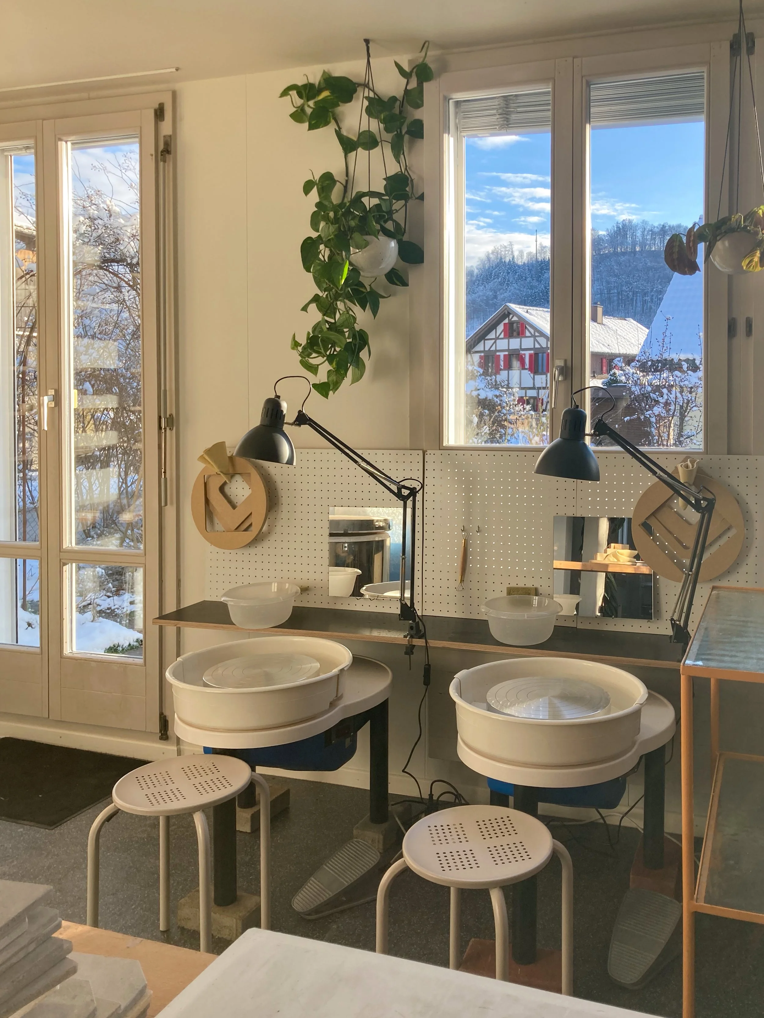 Innenraum einer Töpferei mit Doppel-Topf-Werkbänken, Arbeitslampen, Fenster mit Blick auf Schnee, Haus und Berge, Pflanzen und Stühlen.