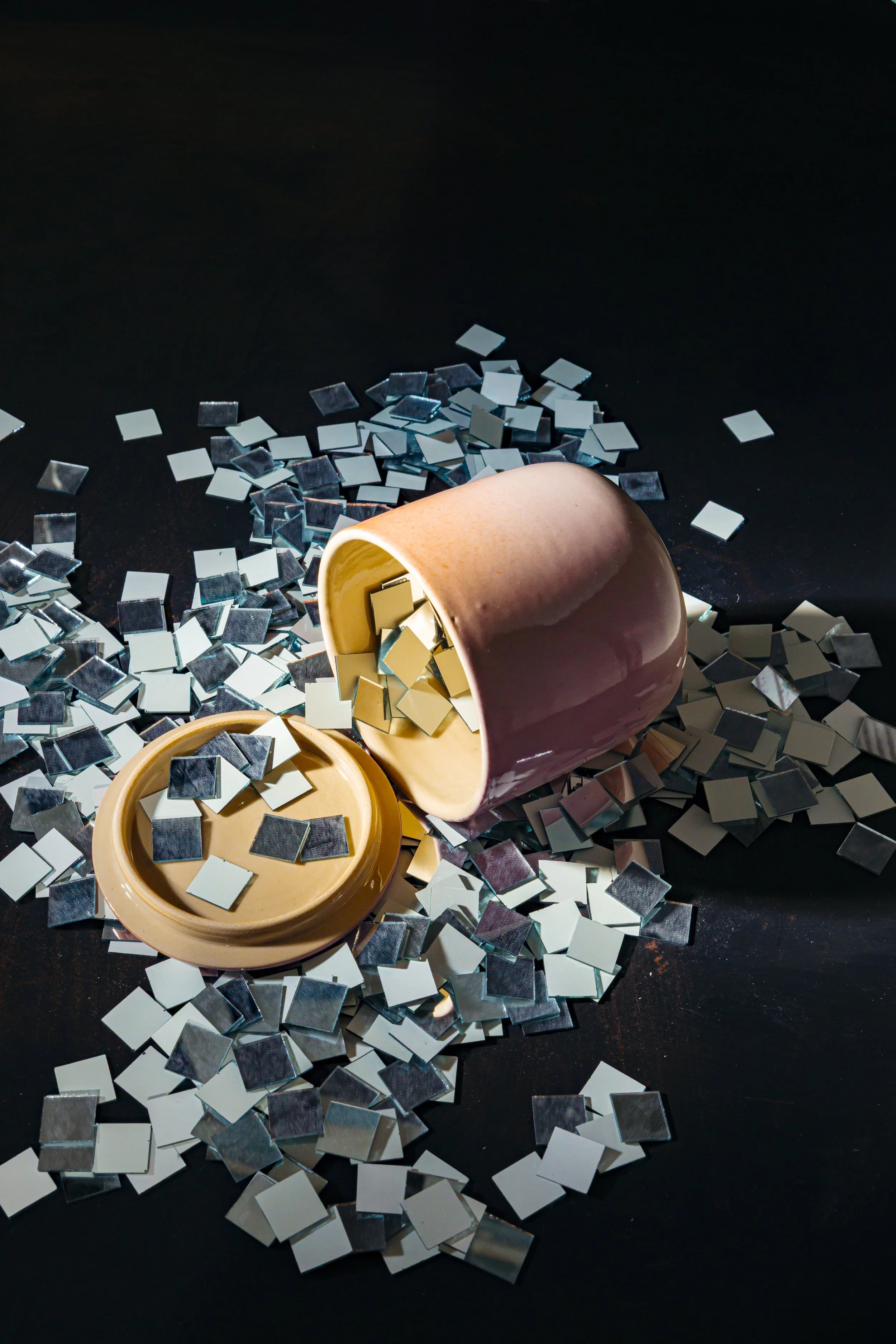 Ein umgedrehter Porzellanbecher mit kleinen quadratischen Spiegelstücken und einem geöffneten Deckel mit weiteren Spiegelstücken, auf einer schwarzen Oberfläche verstreut.