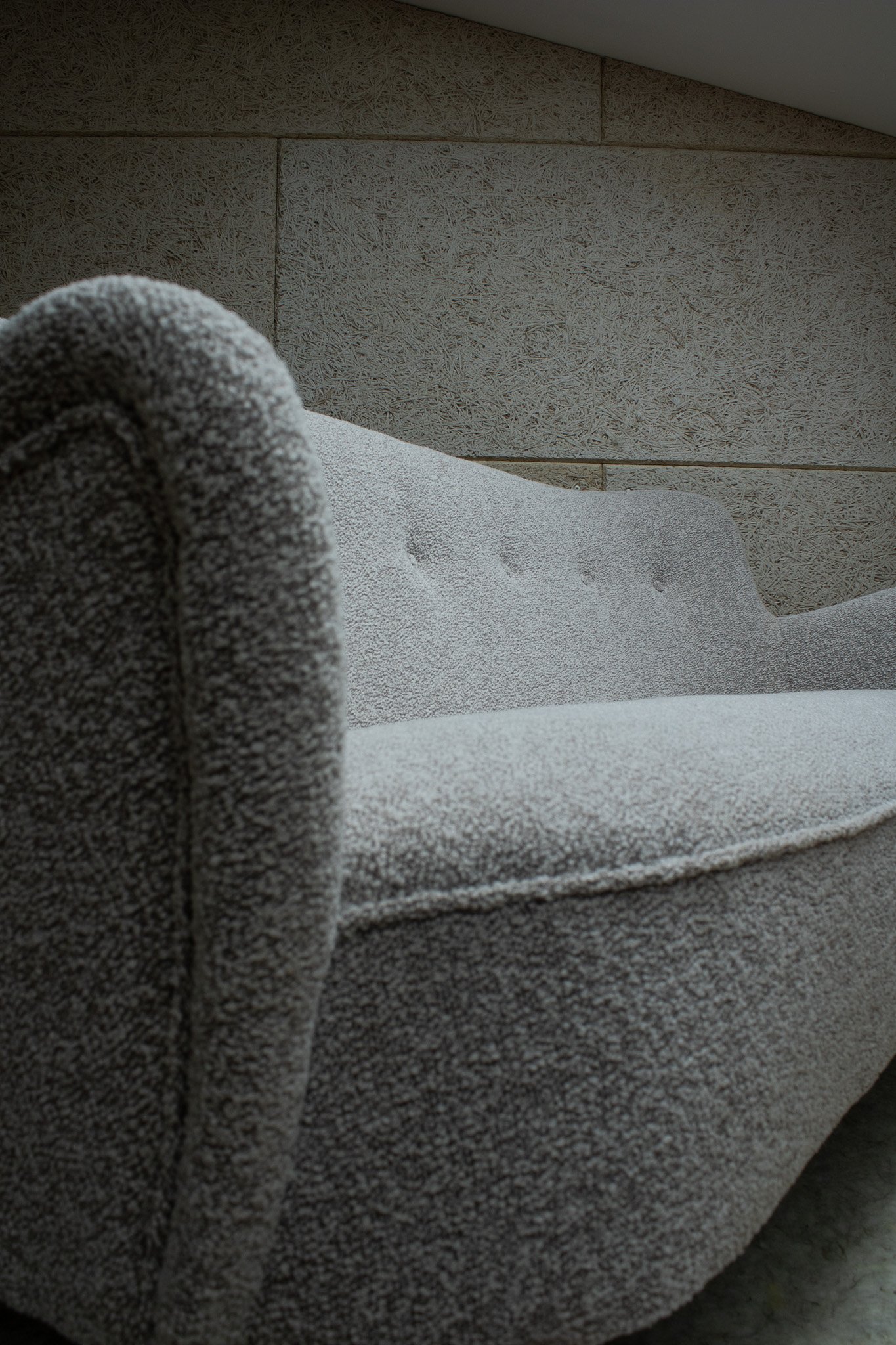 Close-up of a textured, light-colored fabric sofa with a curved armrest, positioned against a wall with textured tiles.