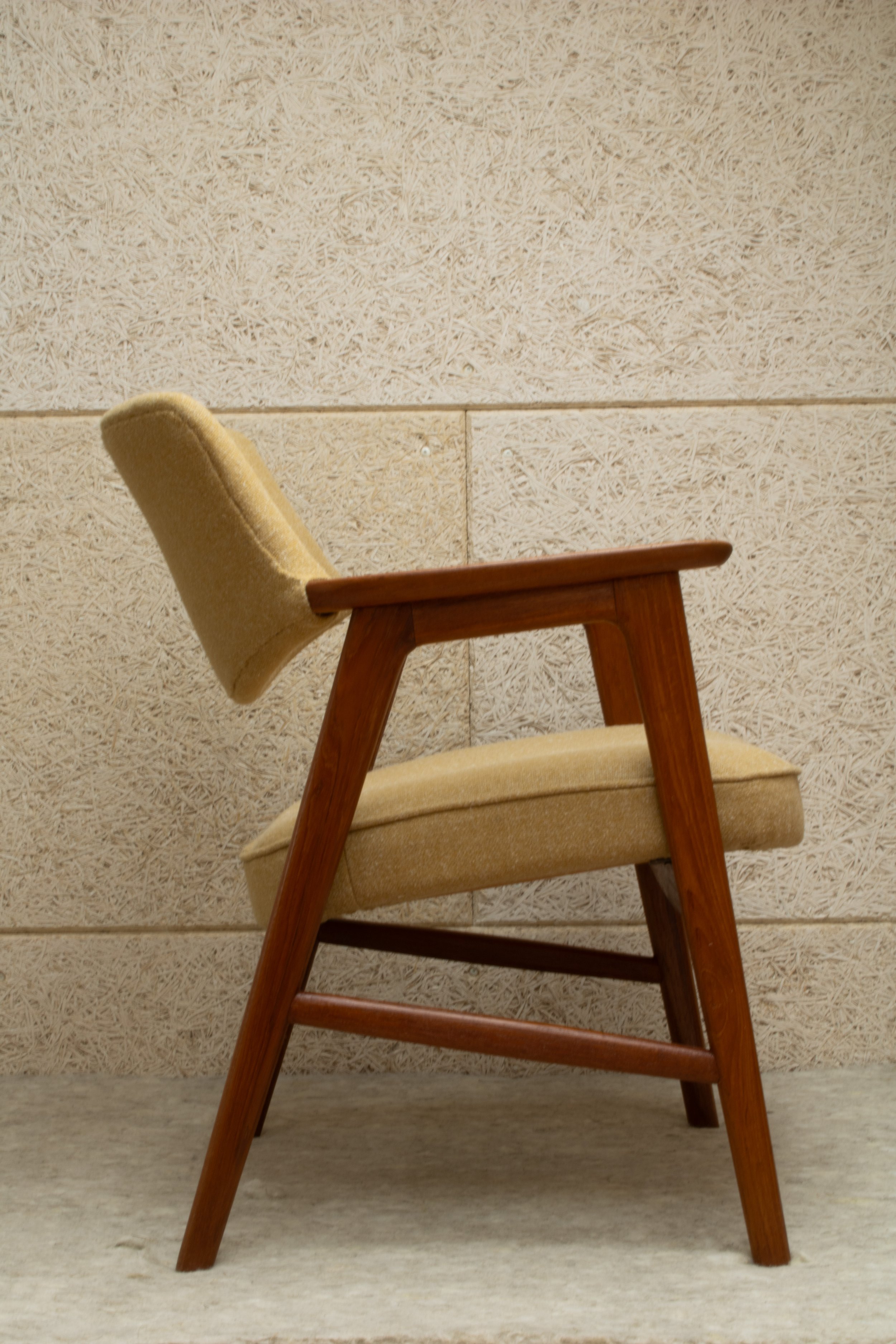 Side view of a mid-century modern armchair with light brown upholstery and wooden frame against a textured beige wall.