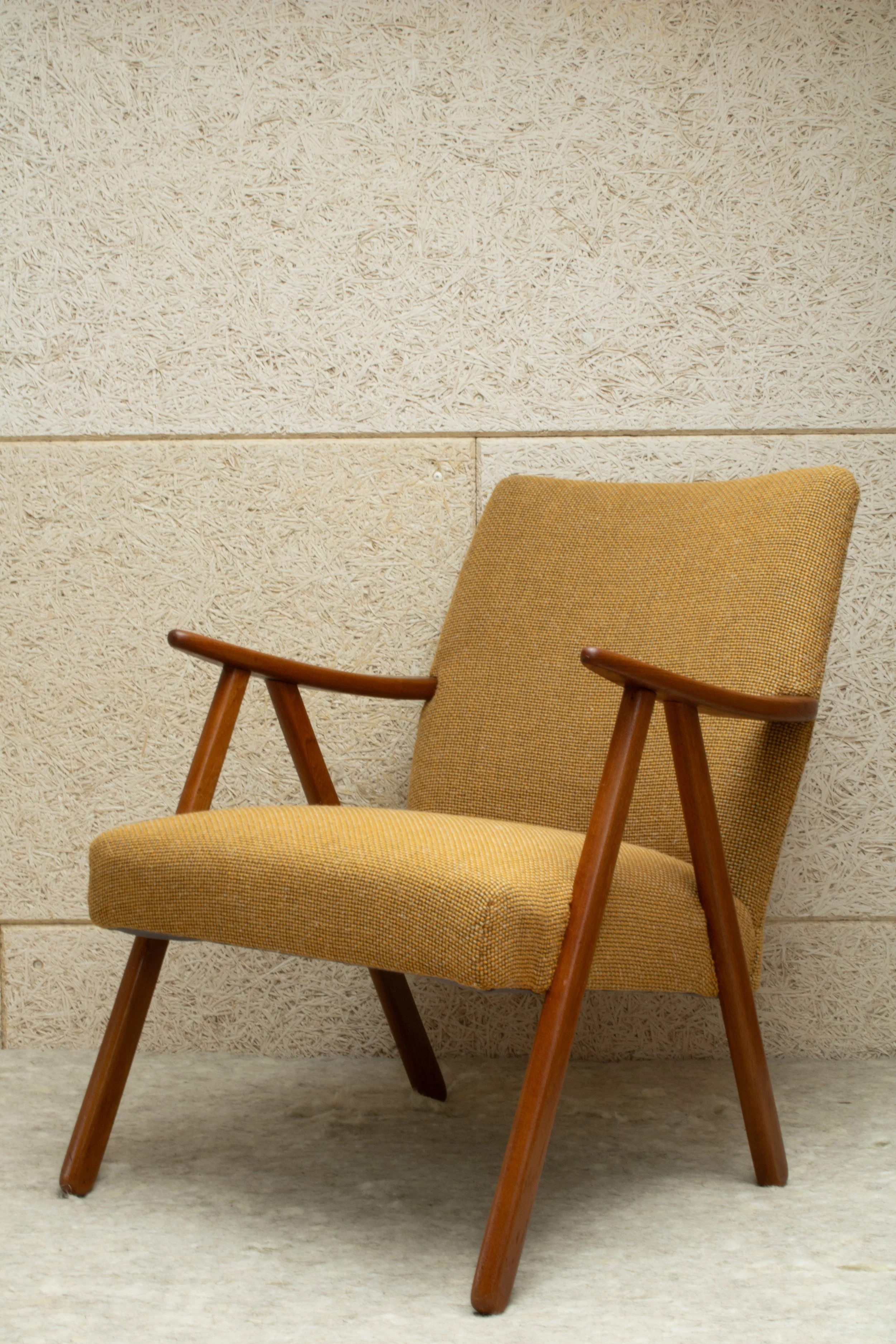 Mid-century modern armchair with wooden arms and legs, upholstered in mustard yellow fabric, placed against a textured beige wall.