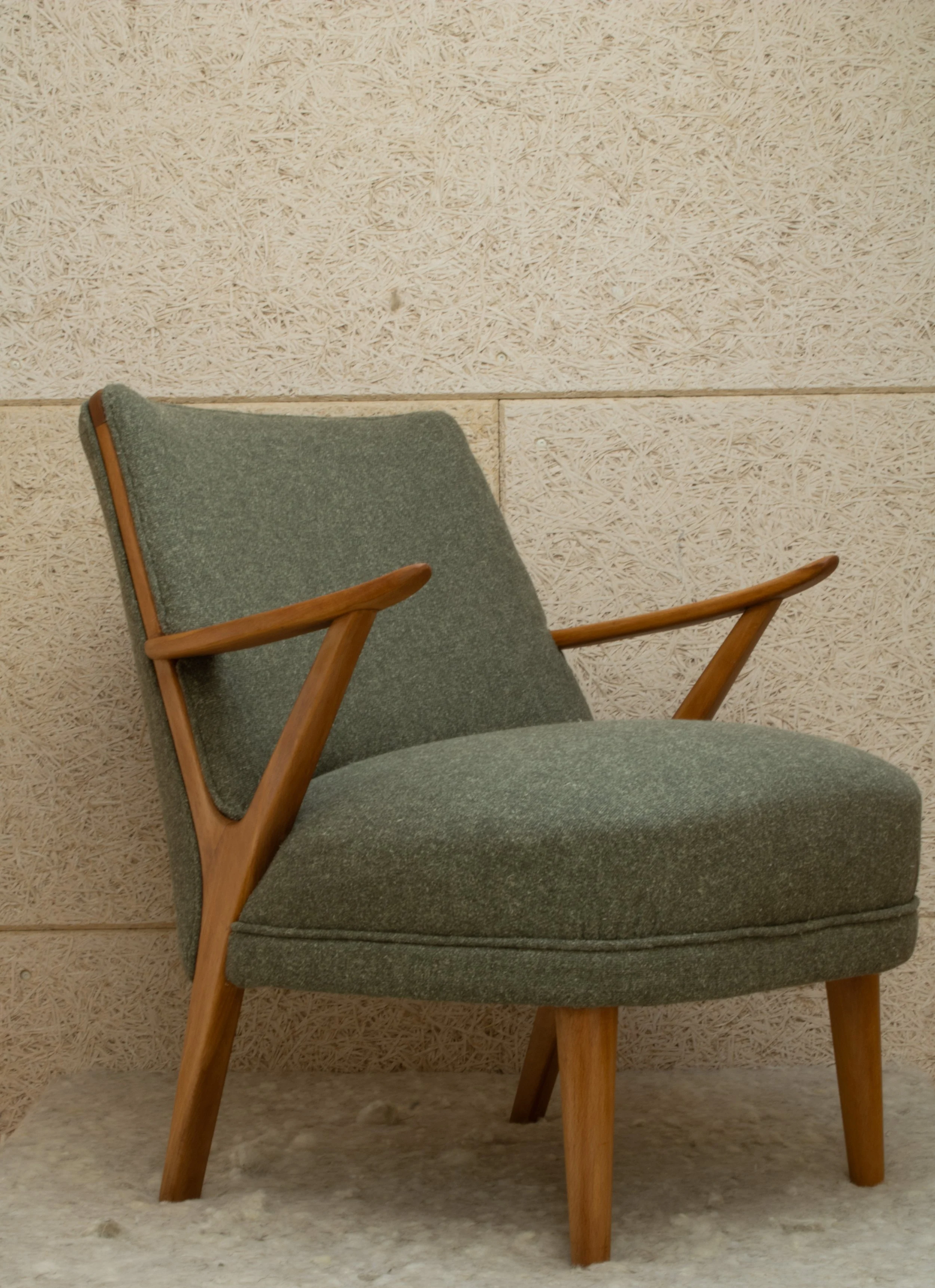 Mid-century modern armchair with green fabric upholstery and wooden frame against a textured beige wall.