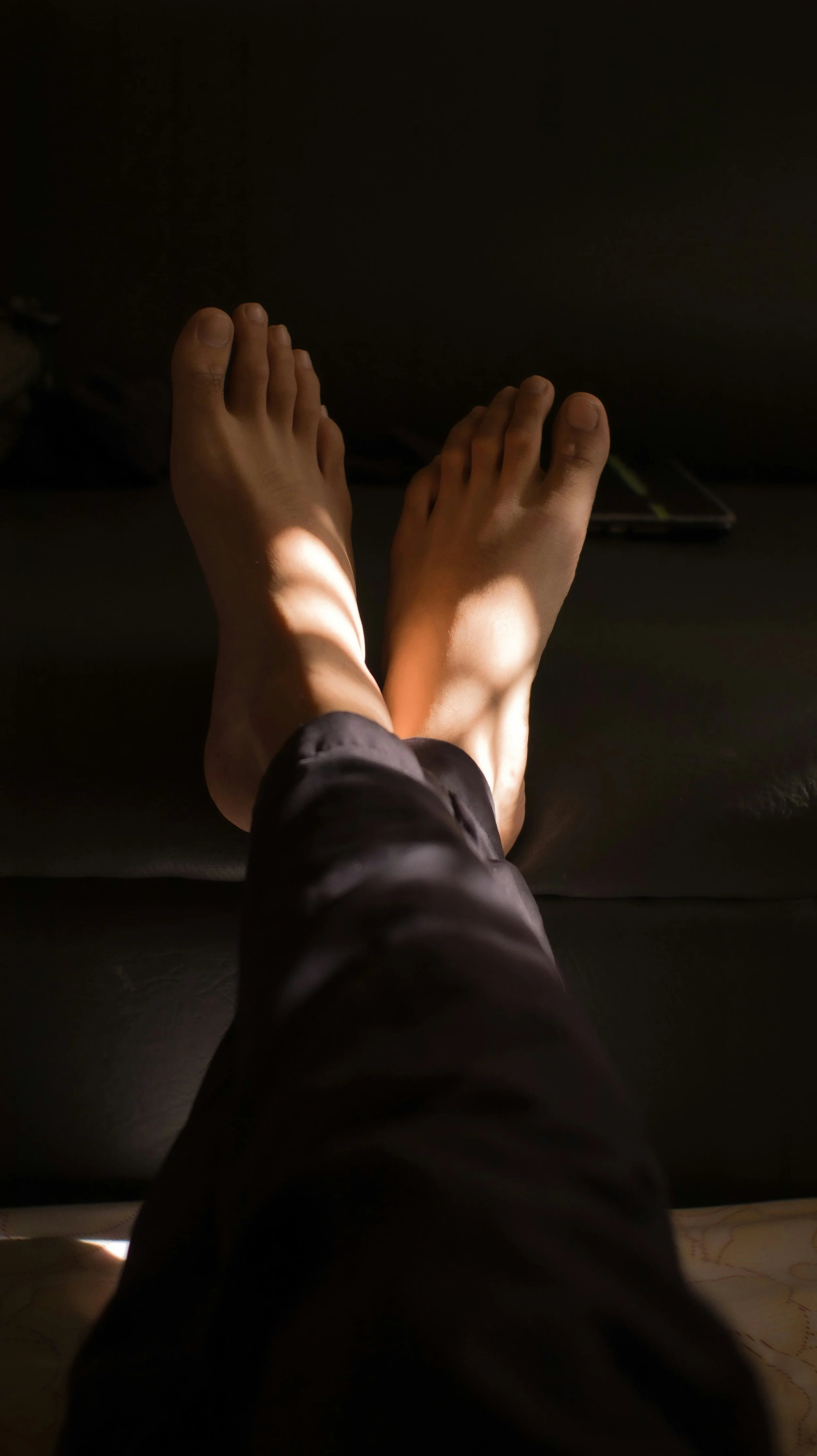 Top-down view of bare feet resting on a black surface with sunlight and shadows falling across them.