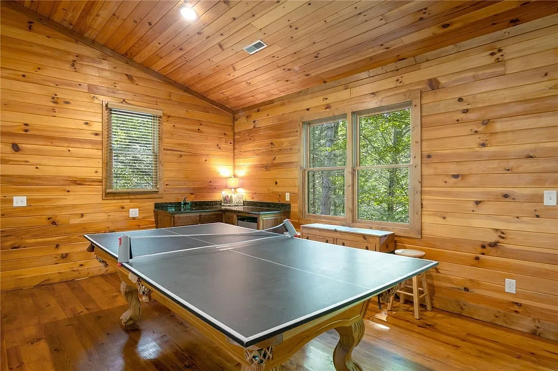 A wooden game room with a ping pong table, a small kitchenette, and large windows overlooking a wooded outdoor area.