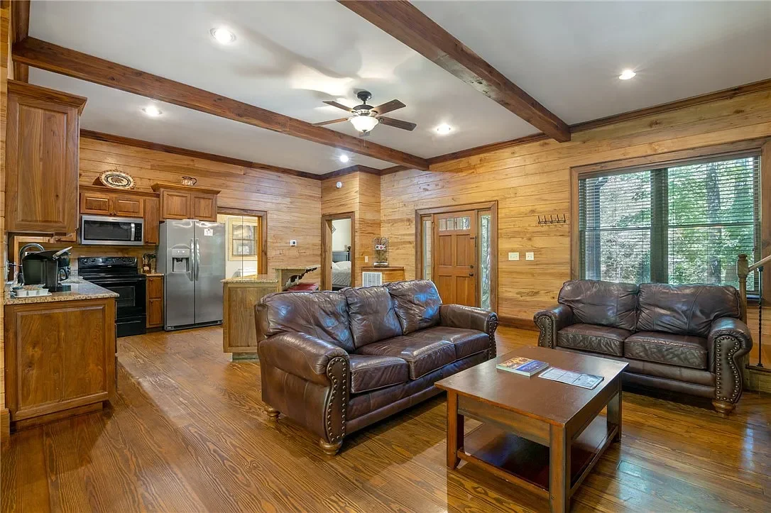 Living room with wooden walls and ceiling, leather couches, a coffee table, large window with blinds, and a kitchen area with stainless steel refrigerator and black appliances.