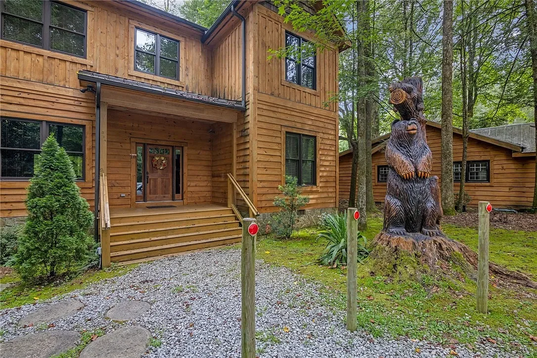 A wooden house with a front porch, surrounded by trees and greenery, featuring a large carved bear statue on a tree stump in the yard.