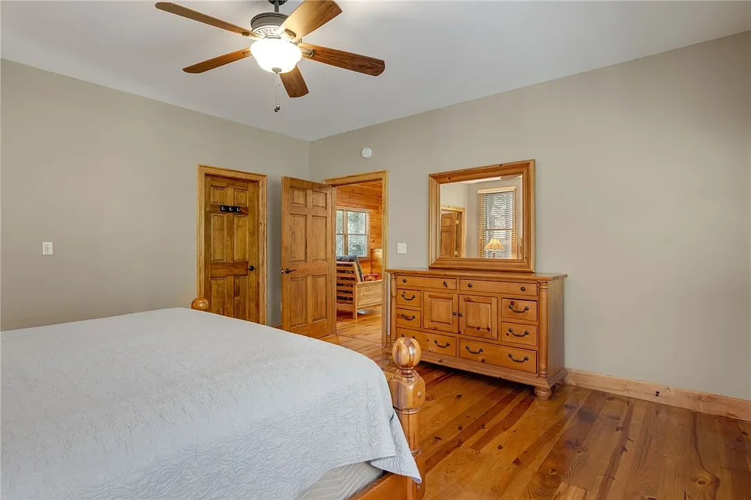 A bedroom with beige walls, hardwood floors, a wooden bed with a white quilt, a wooden dresser with a mirror, and a ceiling fan with wooden blades.