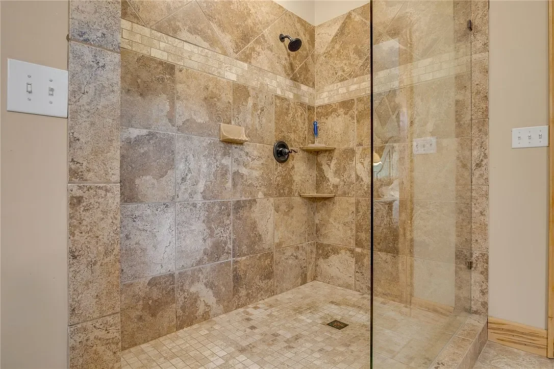A walk-in shower with brown and beige tiled walls and floor, a rain showerhead, a handheld showerhead, and built-in corner shelves.