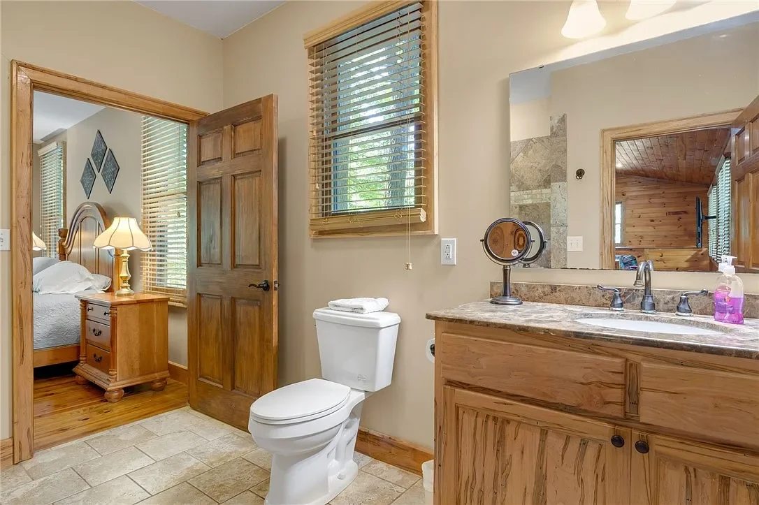 Bathroom with a wooden vanity, a large mirror, and a toilet. There's a window with wooden blinds and a bedroom visible through an open door.