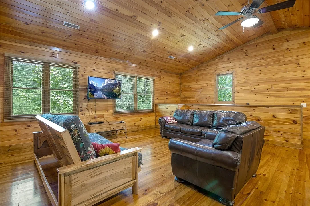 Living room with wooden walls and ceiling, two windows showing trees outside, black leather couches, a wooden futon with cushion, a wall-mounted TV, and a ceiling fan with light.