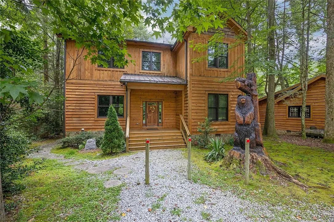 Wooden house in a forested area with a carved bear totem sculpture and a gravel pathway leading to the front steps.