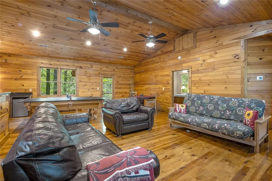 Living room with wooden walls and ceiling, featuring leather and fabric sofas, ceiling fans, windows, and a foosball table.