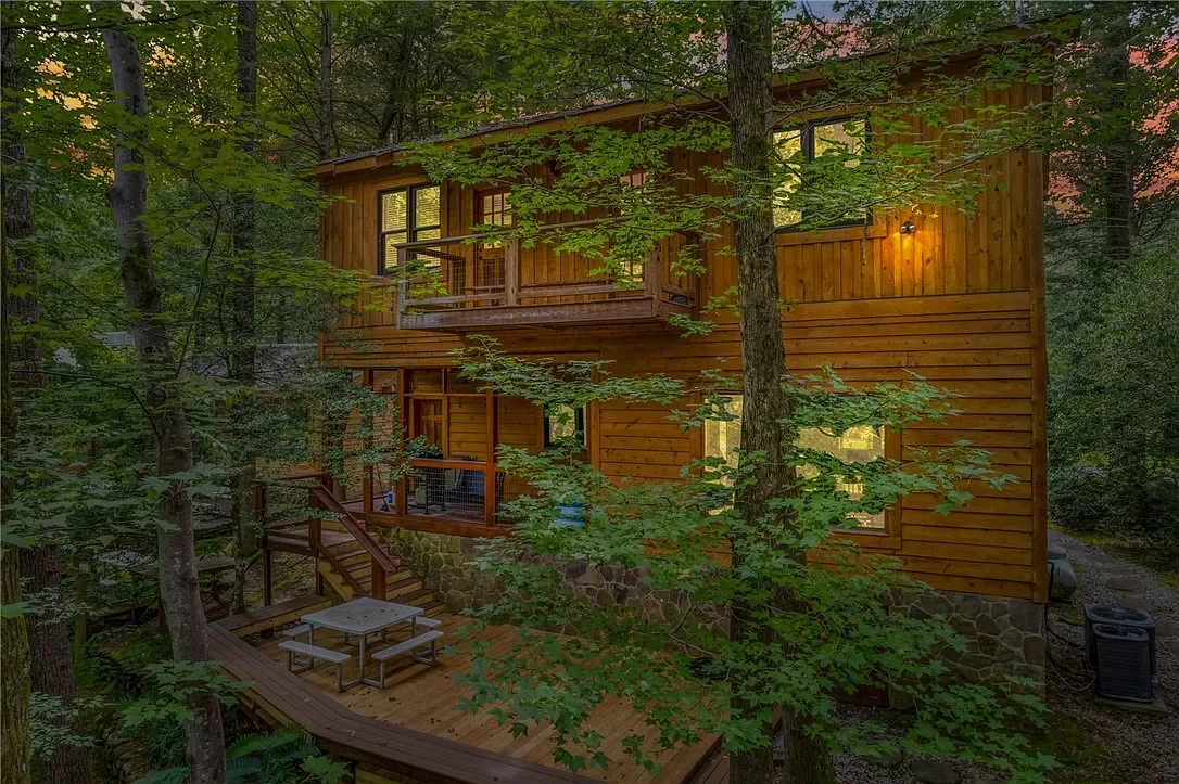 Wooden house surrounded by trees, with outdoor deck, picnic table, and outdoor lighting, in a forest setting at sunset.