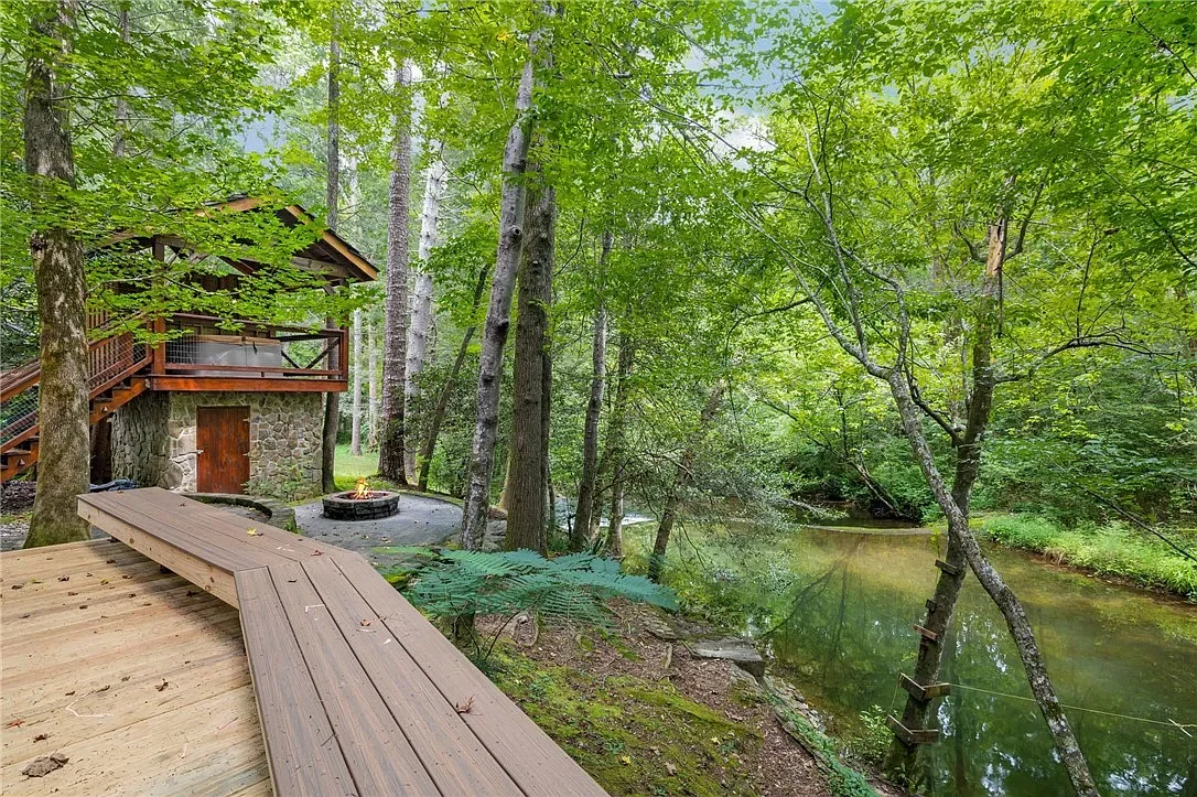 A wooded riverside scene with a wooden deck, a fire pit, and a small building with stone foundation and wooden walls, surrounded by lush green trees.