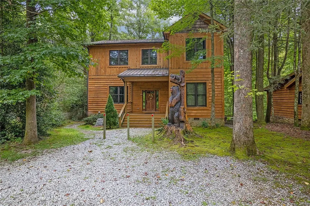 Wooden house surrounded by trees in a forest, with a gravel path leading to the front door, and a carved bear statue on the yard.