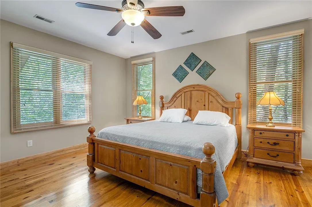 Bedroom with wooden bed and matching nightstands, two lamps, three windows with blinds, ceiling fan, and wall art