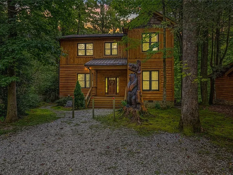 Wooden house surrounded by trees, with a gravel path leading to the front door and a carved bear statue in front of it.