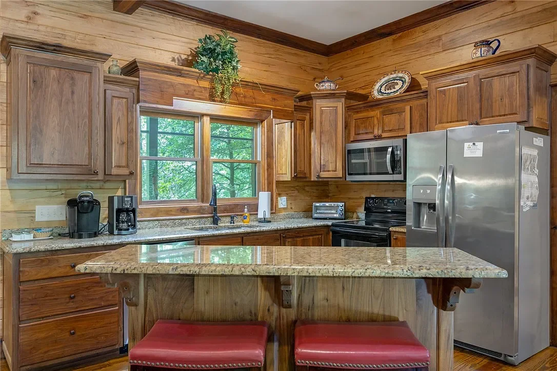 Kitchen with wood cabinets, granite countertops, a window above the sink, a stainless steel refrigerator, microwave, oven, toaster, coffee maker, and coffee machine, and a small breakfast bar with two red stools.