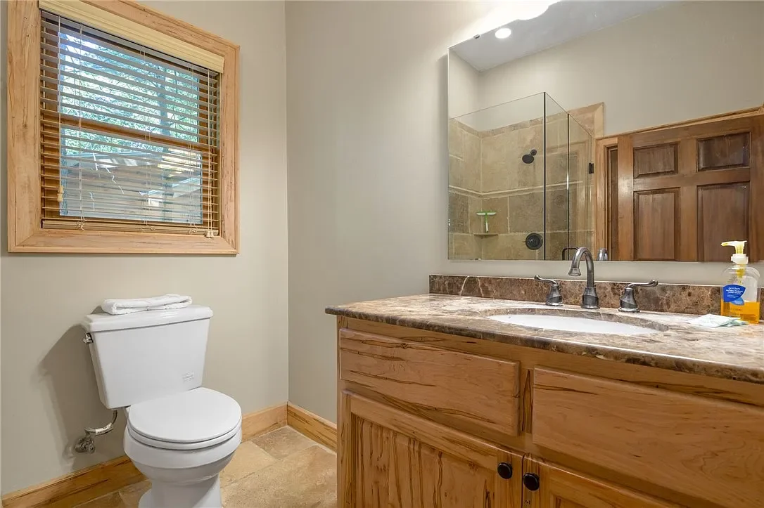 Bathroom with a toilet, wooden framed window with blinds, wooden vanity with a marble countertop, undermount sink, wall-mounted faucet, and glass-enclosed shower with beige tile walls.