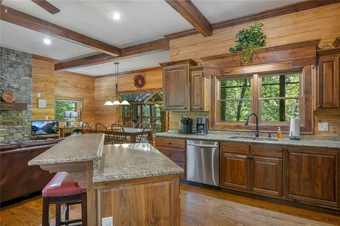 Wood-paneled kitchen with granite countertops, stainless steel dishwasher, large window, and decorative plant above cabinets, open to dining and living area with stone fireplace and wooden beams.