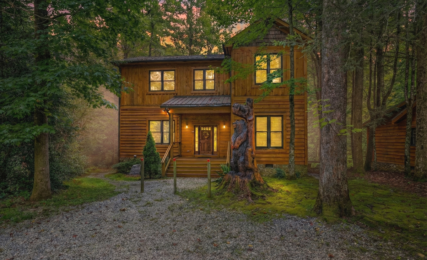 A wooden cabin in a forest at dusk, with lights on inside, surrounded by tall trees and a mossy ground.
