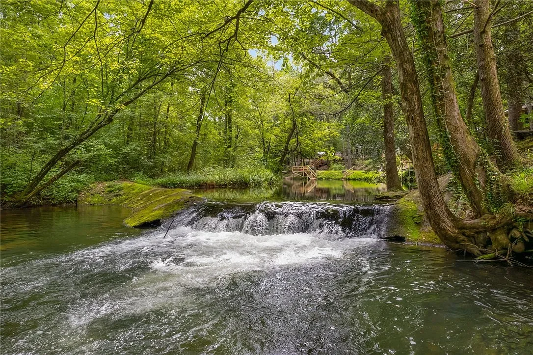 A peaceful creek flowing through a green forest with trees and grass, with a small waterfall in the foreground and a wooden walkway and house in the background.
