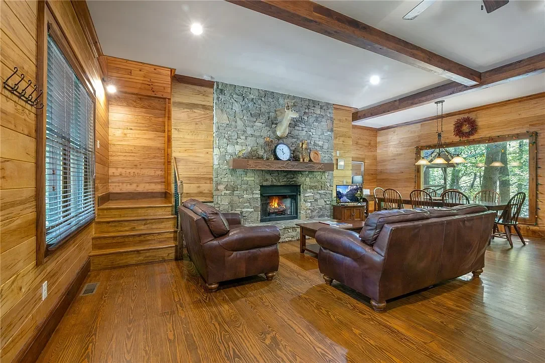 Living room with wood-paneled walls, hardwood flooring, a stone fireplace with a mantel, two leather sofas, a wooden coffee table, a dining area with a table and six chairs, large windows with curtains, and a wood ceiling with exposed beams.