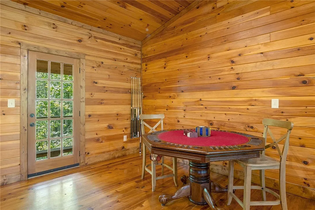 Wood-paneled dining area with a round table covered with a red tablecloth and two chairs, next to a door with glass panes and a rack holding several pool cues.