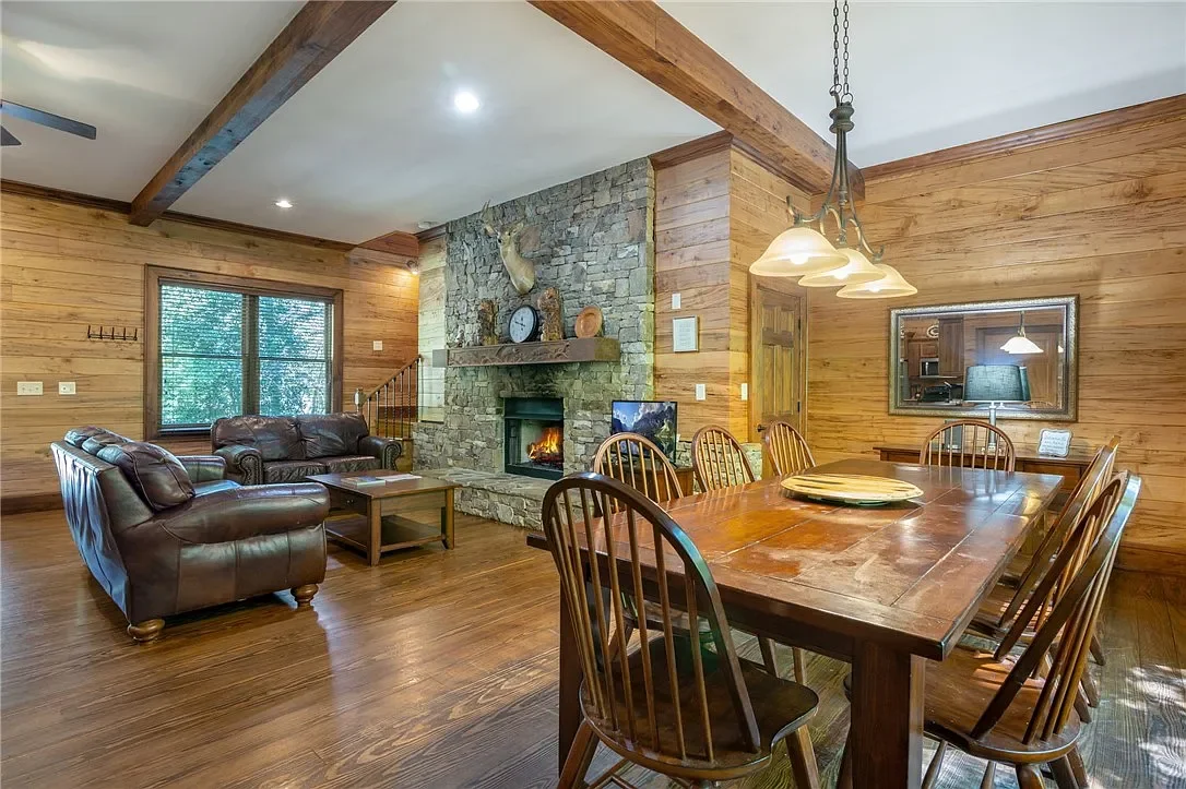 Interior of a cozy living and dining area with wood-paneled walls, a stone fireplace, leather sofas, a large wooden dining table, and a window with a view of trees.