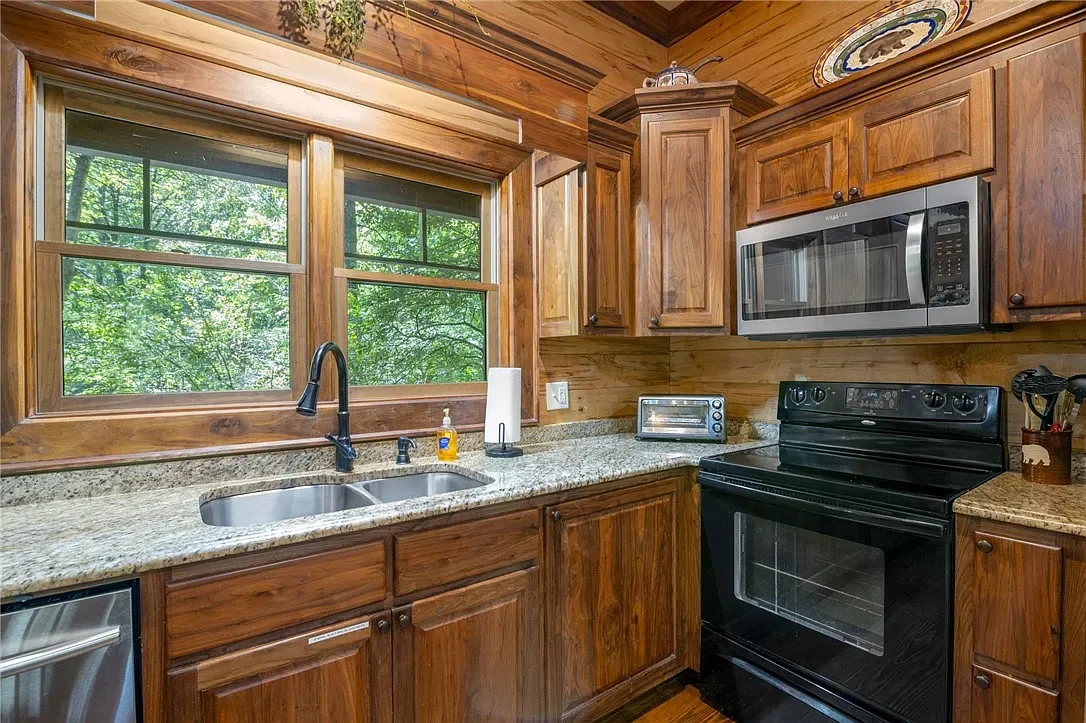Kitchen with wood cabinets, granite countertops, a double sink, and a large window showing green trees outside. Includes a microwave, small toaster oven, black stove, and various kitchen essentials.