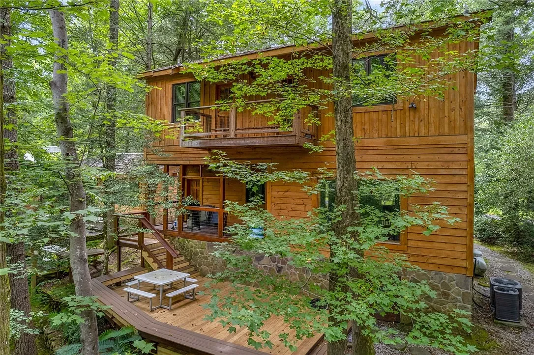 Wooden house with a stone foundation surrounded by trees, featuring a deck with outdoor furniture and a balcony.