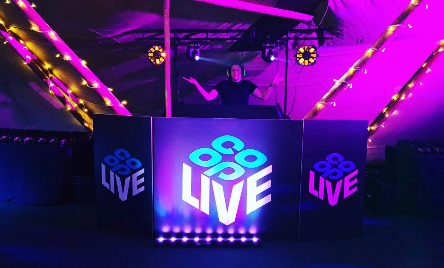 A DJ wearing headphones is standing behind a DJ booth with purple and blue lighting, displaying the word 'LIVE' and a logo with circles and the text 'O O O' at a night event.