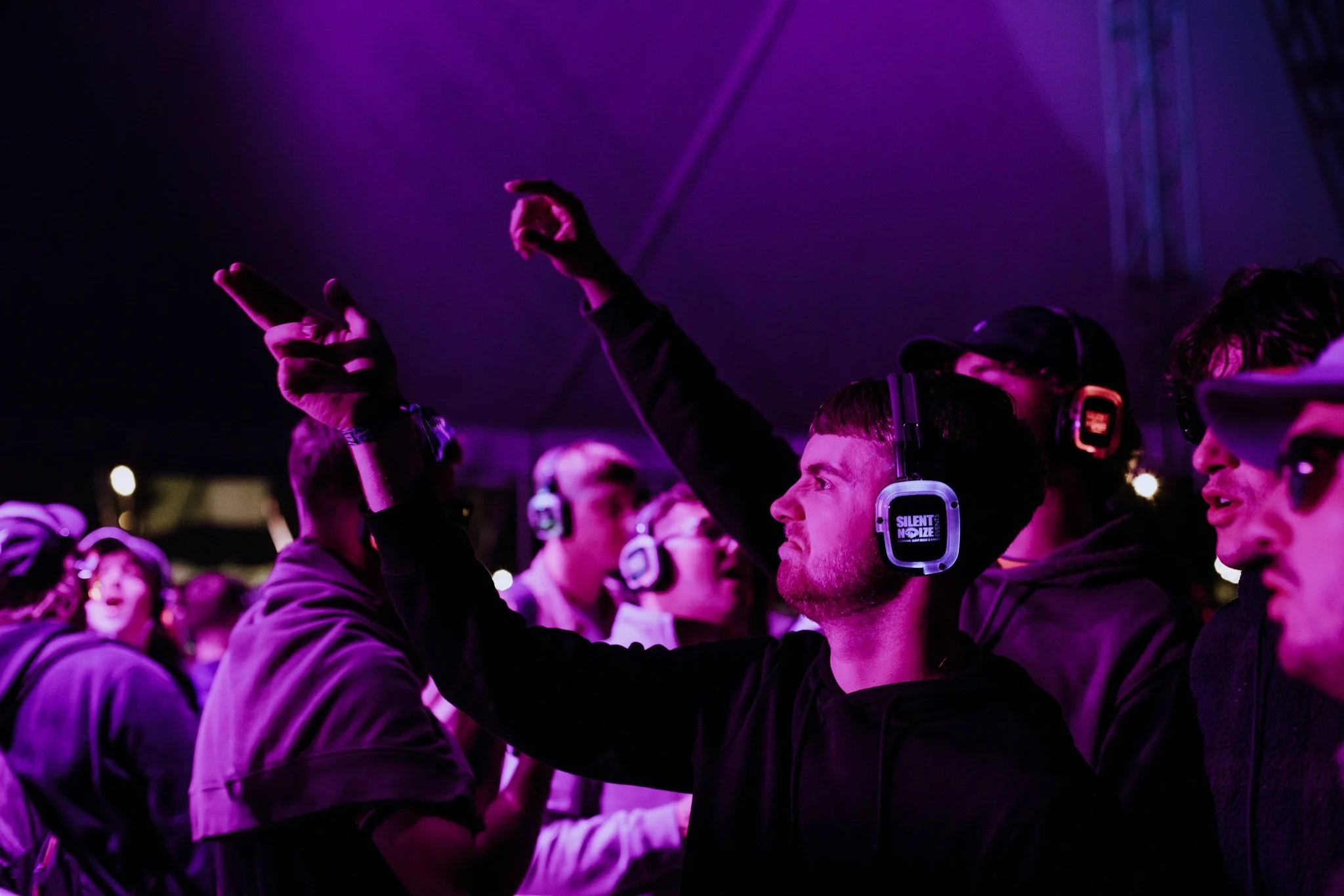 Young man at a music event, wearing headphones, pointing upwards and looking engaged, with purple lighting and other people in the background.
