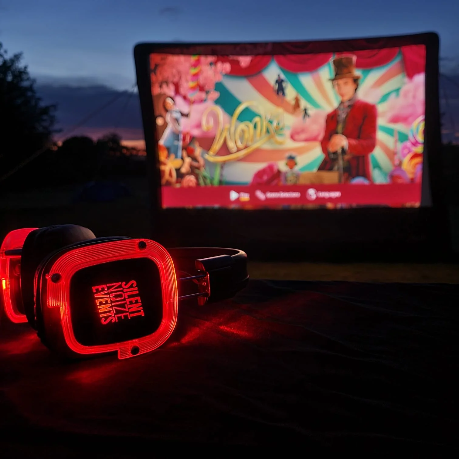 A pair of red guitar pick-shaped headphones with the text 'SILENT NOIZE EVENTS' on the earpiece, placed on a dark surface outside during twilight. In the background, a large outdoor screen displays a colorful image with a person in a red jacket and a bowler hat, and the sky is darkening.