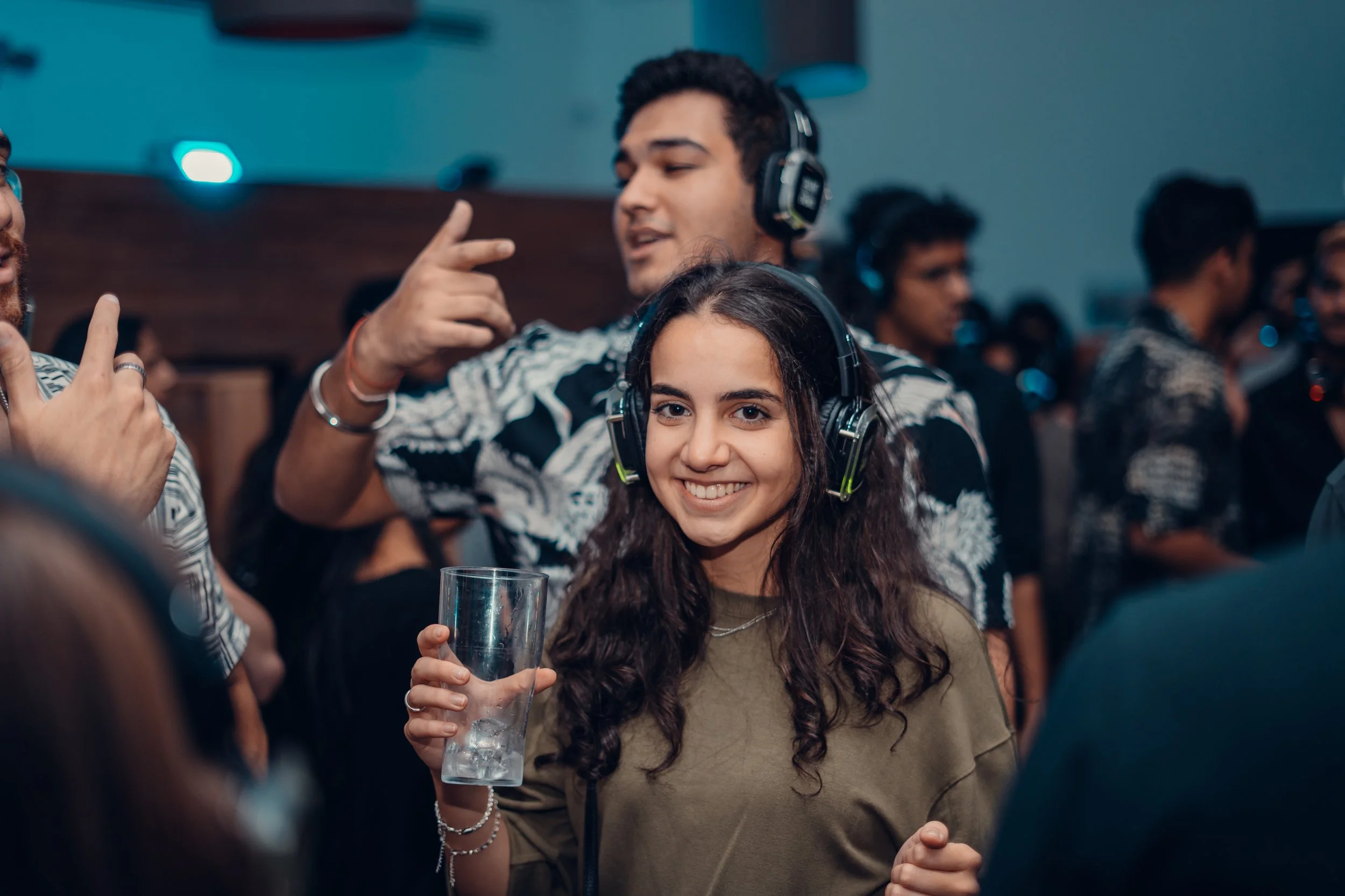 Smiling young woman wearing headphones holding a glass of drink at a lively party or event with people dancing in the background.