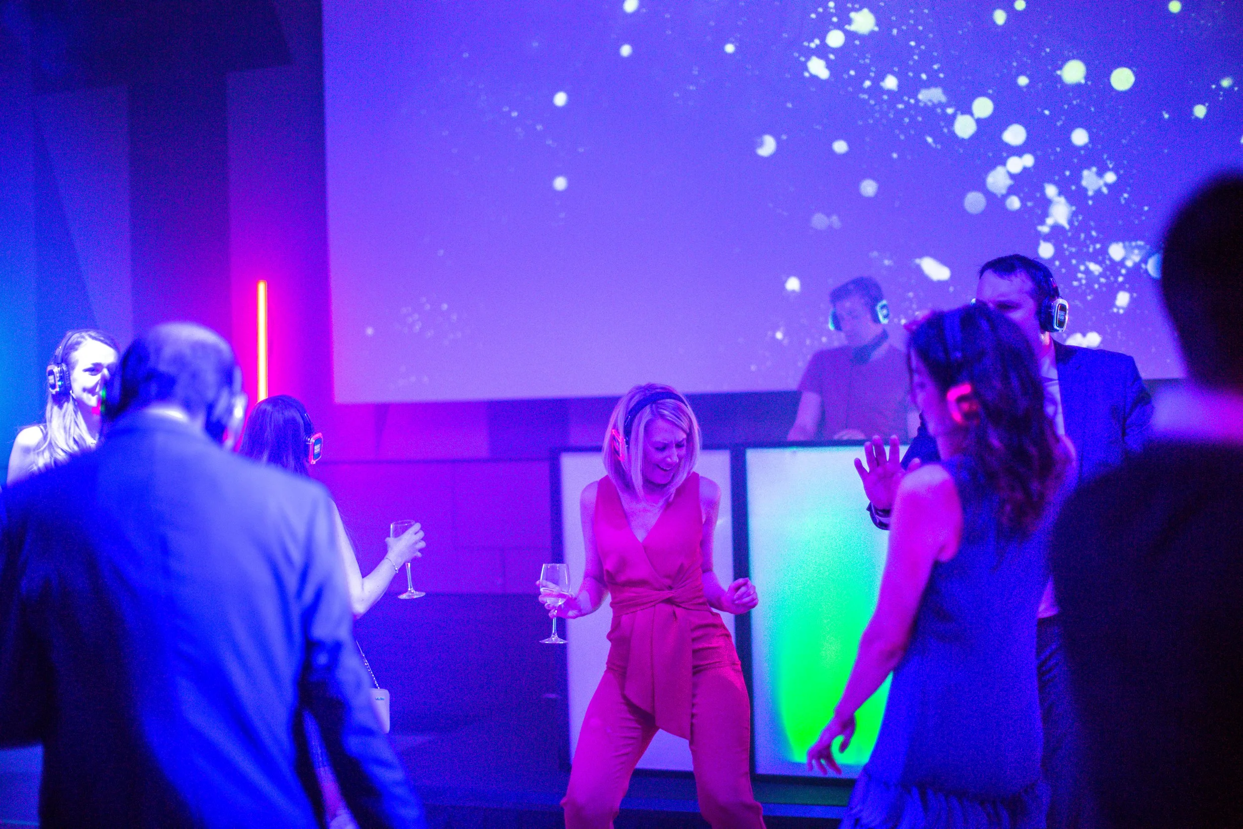 People dancing and enjoying music at a nightclub with colorful lighting and a large digital screen in the background.