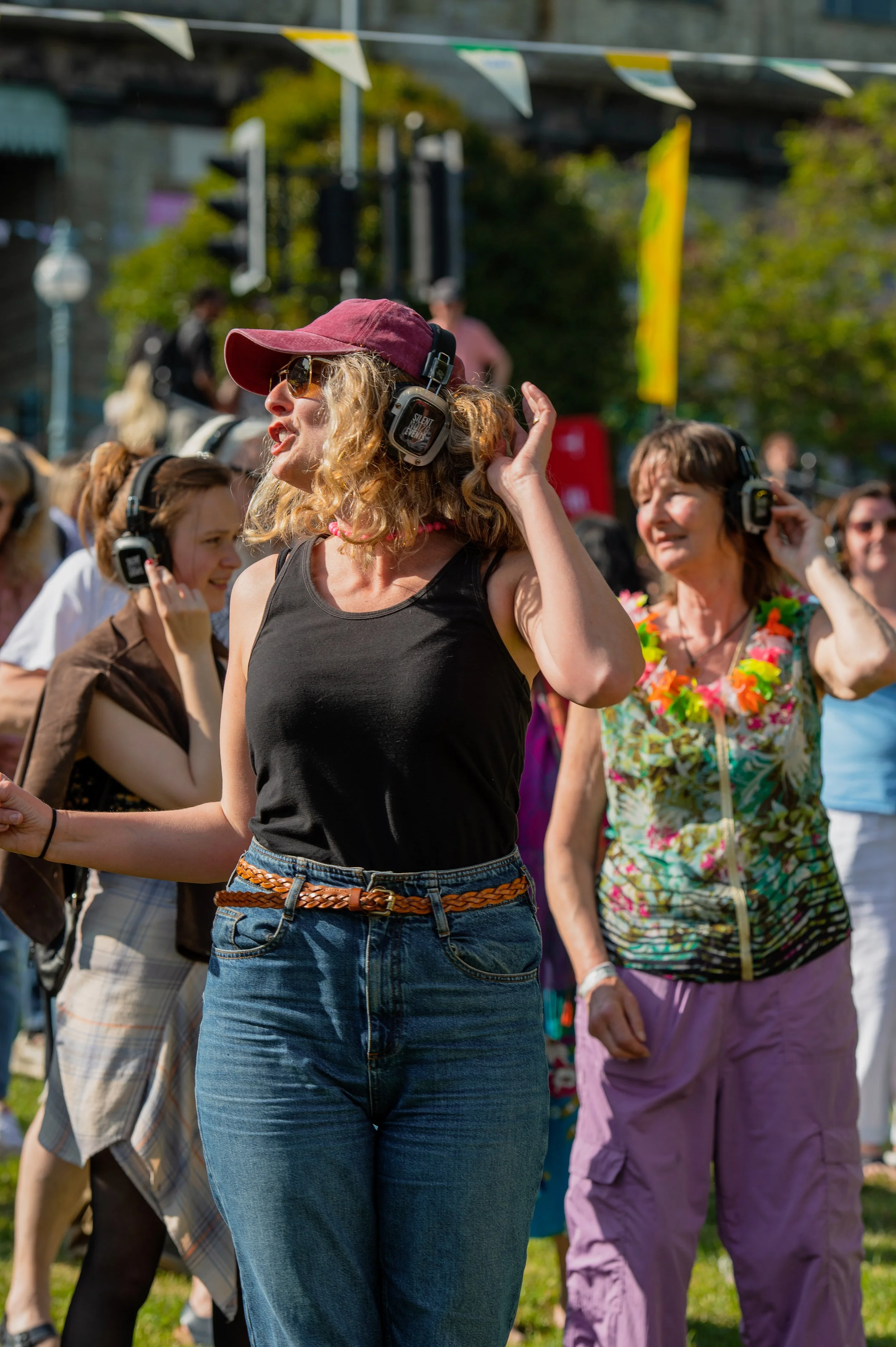 Group of people at an outdoor event, some wearing headphones, with a woman in the foreground wearing sunglasses, a maroon baseball cap, a black sleeveless shirt, and blue jeans, talking or singing.