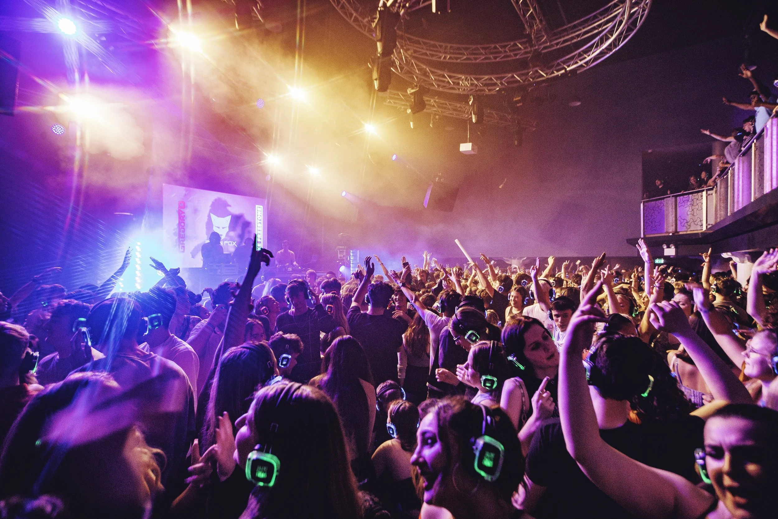 Crowd of people dancing and enjoying music at a nightclub with colorful lights and a DJ on stage.