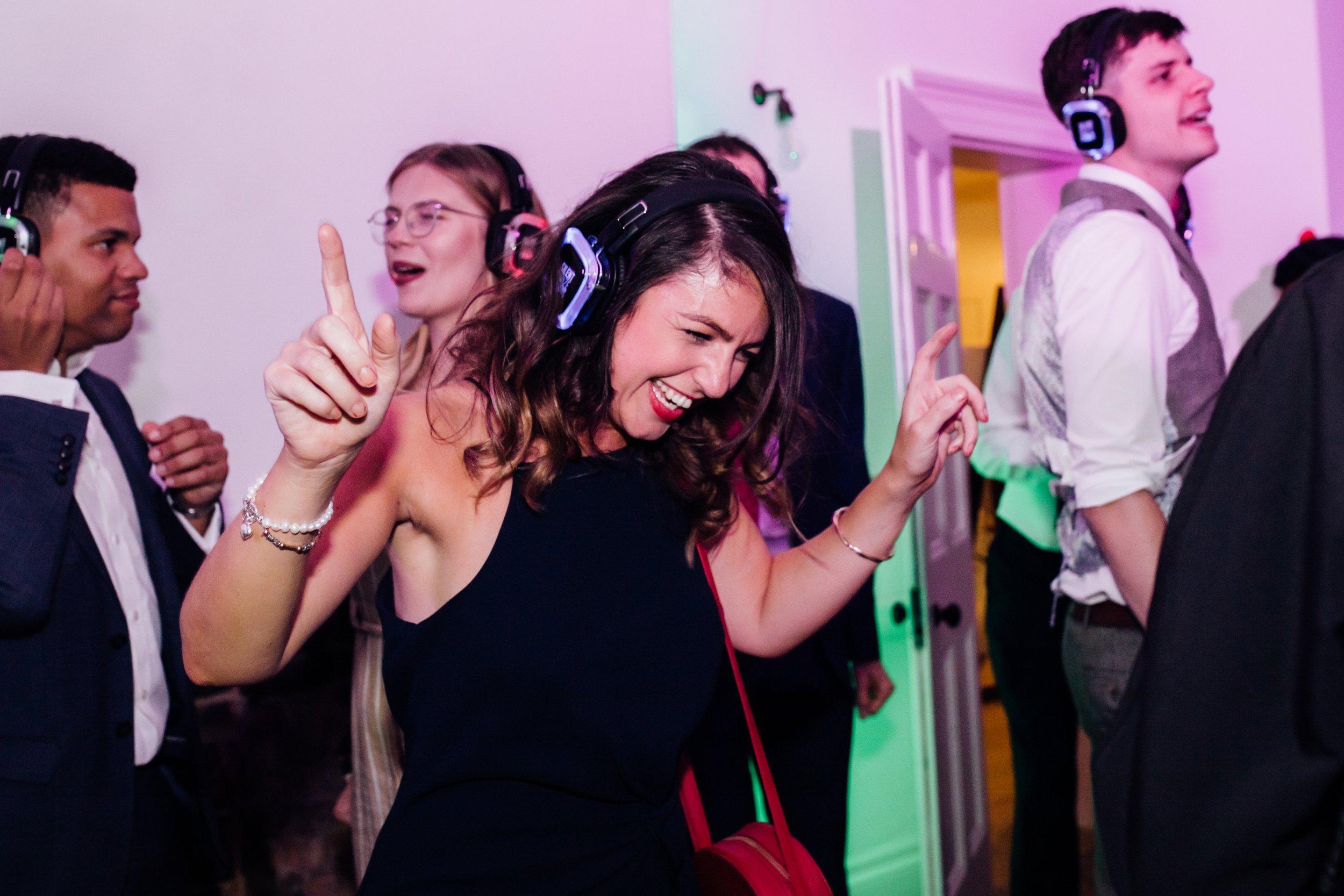 A woman dancing and smiling at a party wearing headphones, surrounded by other people also enjoying music.