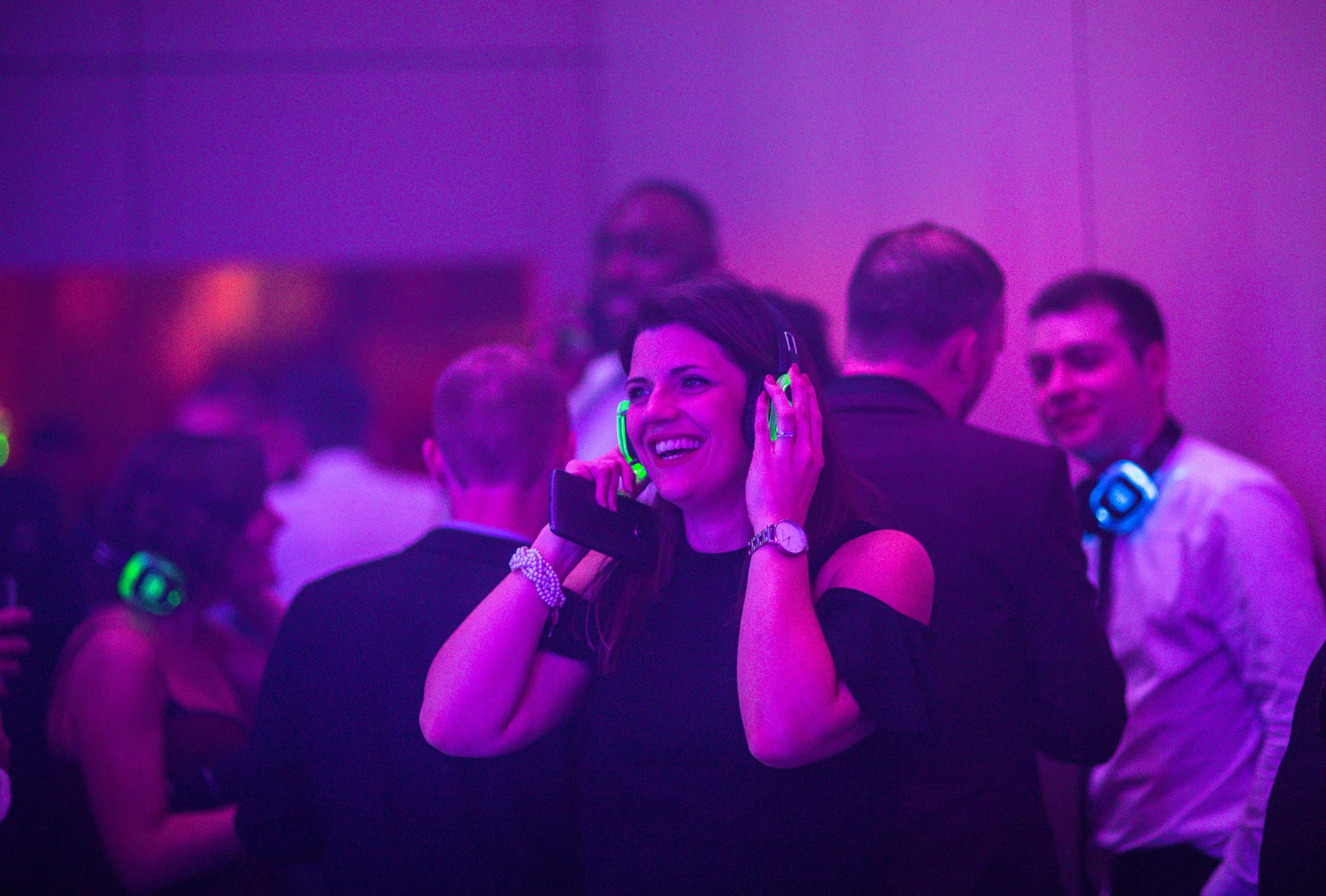 Group of people at a party with colorful lighting, some wearing headphones, smiling and chatting.