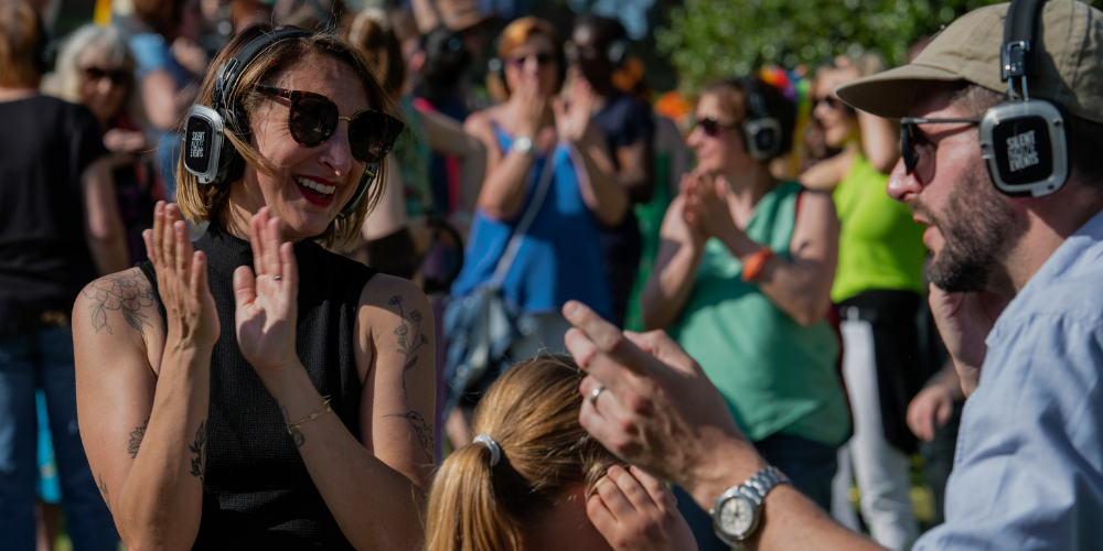 A woman with sunglasses and tattoos smiling and clapping at an outdoor event, with a man wearing headphones and sunglasses holding a phone or camera, surrounded by other people in the background.